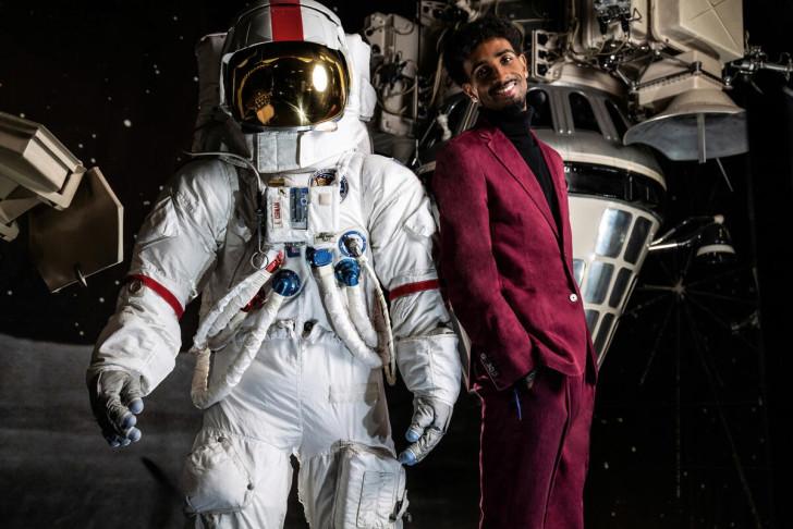 Allan Petre, en costume, est adossé à une combinaison spatiale